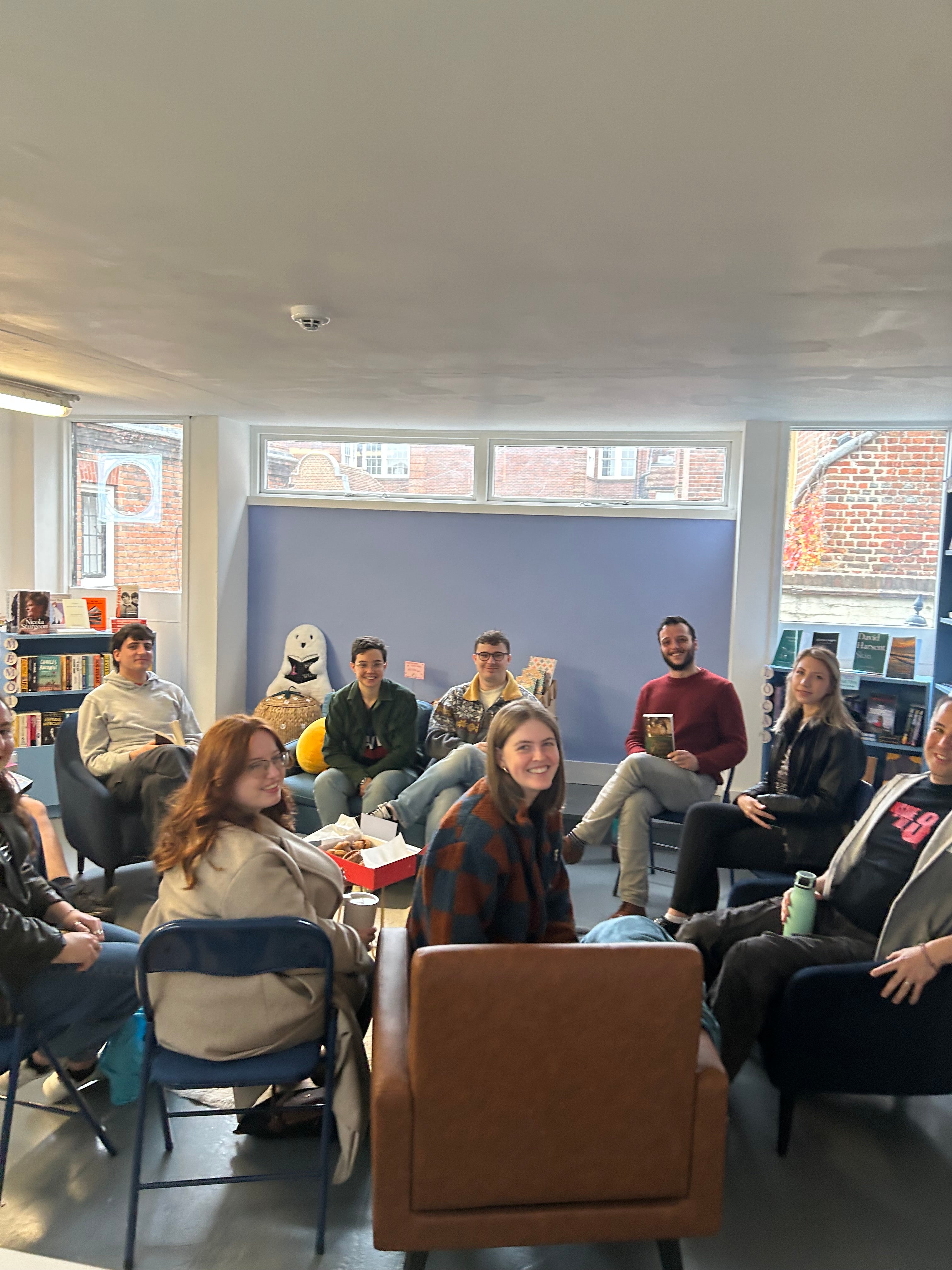 A group of people sitting in a casual indoor meeting or workshop setting, arranged in a circle around a table in a modern room with large windows and a blue accent wall.